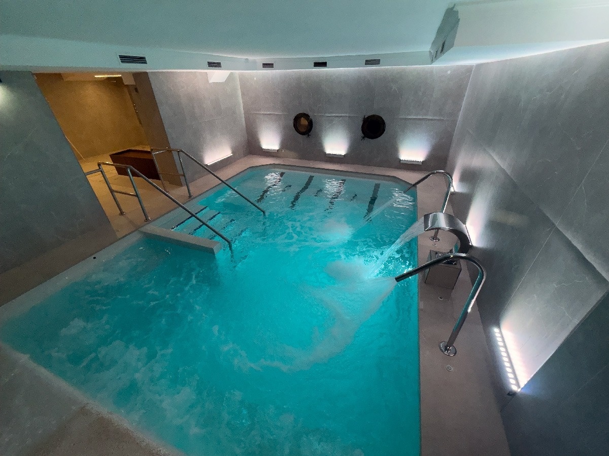 Un moderno spa interior presenta una piscina de agua turquesa con chorros de agua y una iluminación ambiental sofisticada.
