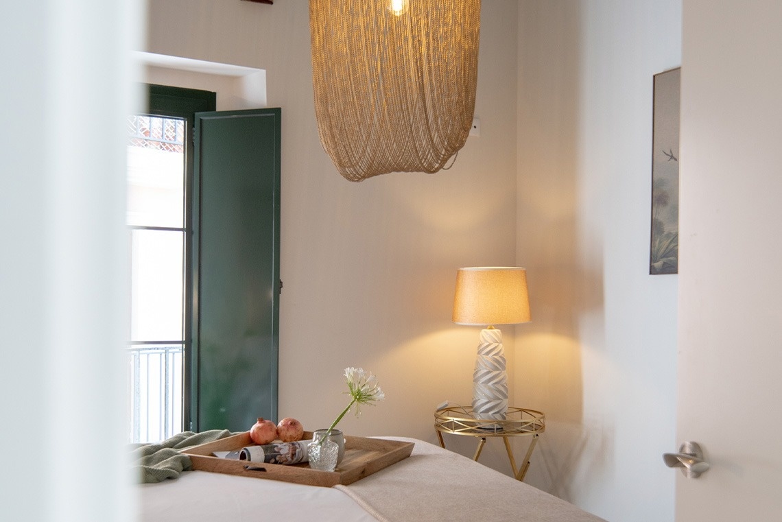 Una habitación bien iluminada presenta una cama con una bandeja de fruta y flores, una lámpara de mesilla encendida, una ventana con postigos verdes y una distintiva lámpara de techo de cadenas doradas.