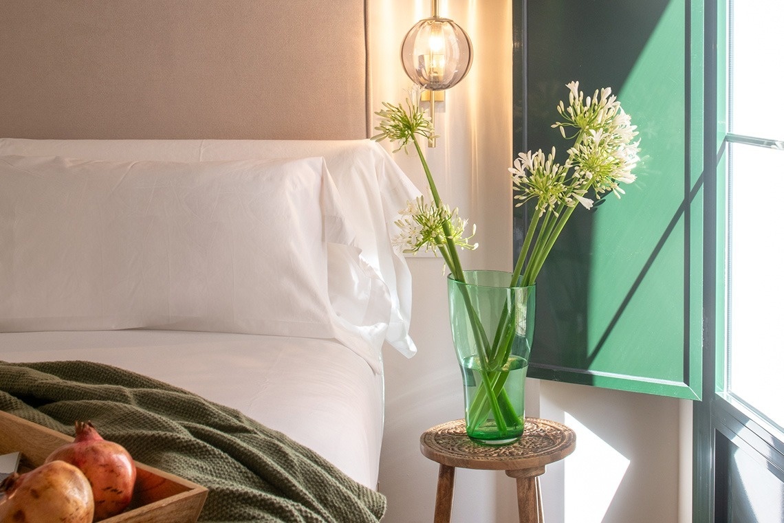 Un dormitorio luminoso presenta una cama con sábanas blancas y una manta verde, un jarrón de flores blancas sobre una mesita de noche, un plato con granadas y un panel de pared verde oscuro, todo ello bañado por la luz natural que entra por una ventana.