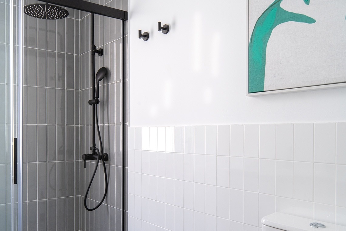 Un moderno cuarto de baño presenta una ducha con azulejos grises verticales y grifería negra, junto a una pared blanca con azulejos blancos y un cuadro abstracto verde.