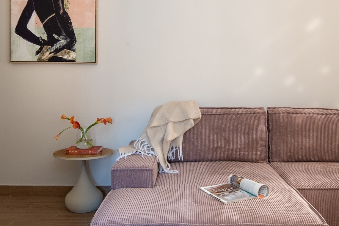 Una acogedora sala de estar presenta un sofá seccional de pana malva con una manta, una mesa lateral con lirios de cala naranjas y un arte abstracto en la pared clara.