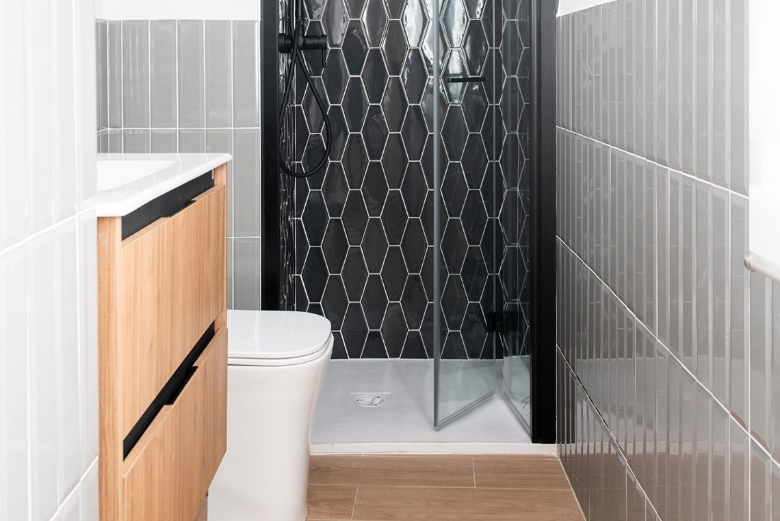 Un baño moderno presenta un inodoro junto a un mueble de madera, una ducha con azulejos hexagonales negros, paredes con azulejos rectangulares grises y blancos, y un suelo de baldosas de madera.