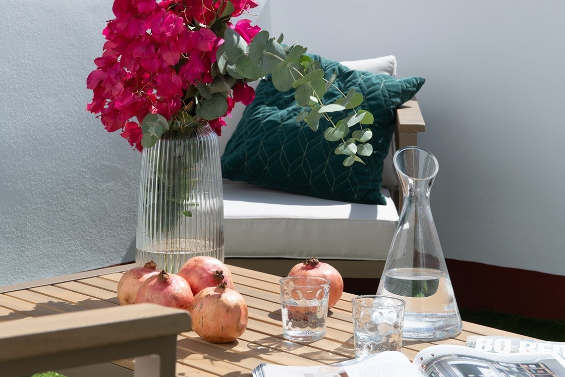 Una mesa de madera al aire libre con un jarrón de flores fucsias y ramas de eucalipto, granadas, vasos y una jarra de agua, y un cojín verde en una silla de fondo.