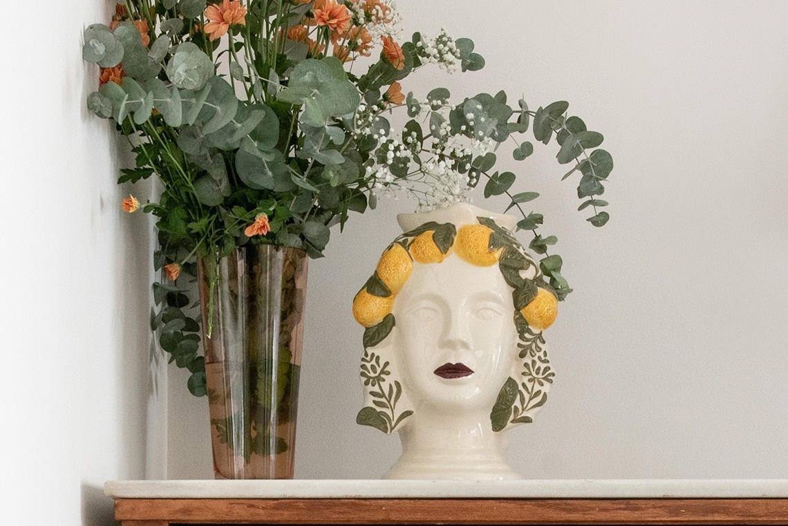 Sobre una repisa, un jarrón con flores de eucalipto y anaranjadas acompaña a un busto de cerámica adornado con limones y follaje.