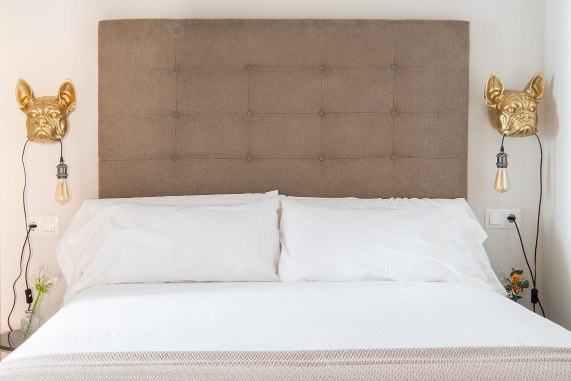 Una cama con un cabecero marrón capitoné está flanqueada por dos lámparas de pared doradas con forma de bulldog y tiene ropa de cama blanca.