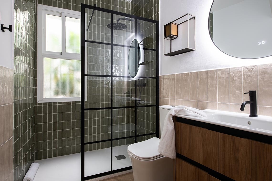 Un baño moderno presenta una ducha con mampara de cristal enmarcada en negro, azulejos verde oscuro y beige, un inodoro, un lavabo con mueble de madera y grifo negro, y un espejo redondo iluminado.