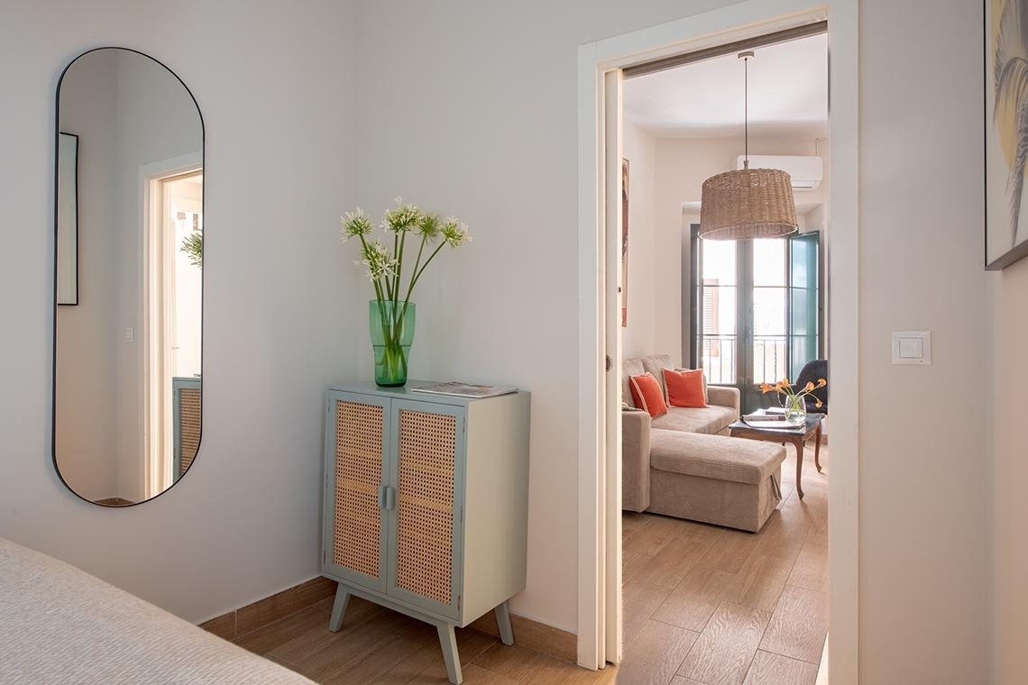 La imagen muestra una zona de entrada con un espejo vertical y un mueble con un jarrón de flores, que se abre a una sala de estar con un sofá, cojines naranjas y un balcón.