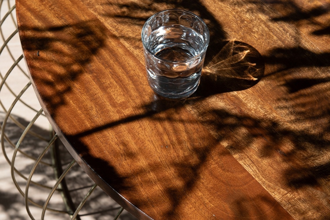 Un vaso de agua descansa sobre una mesa de madera, bajo la luz del sol que dibuja intrincadas sombras de follaje.