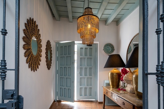Una entrada luminosa y decorada con una puerta azul abierta, un candelabro colgante, espejos dorados en la pared y una mesa consola con lámparas.