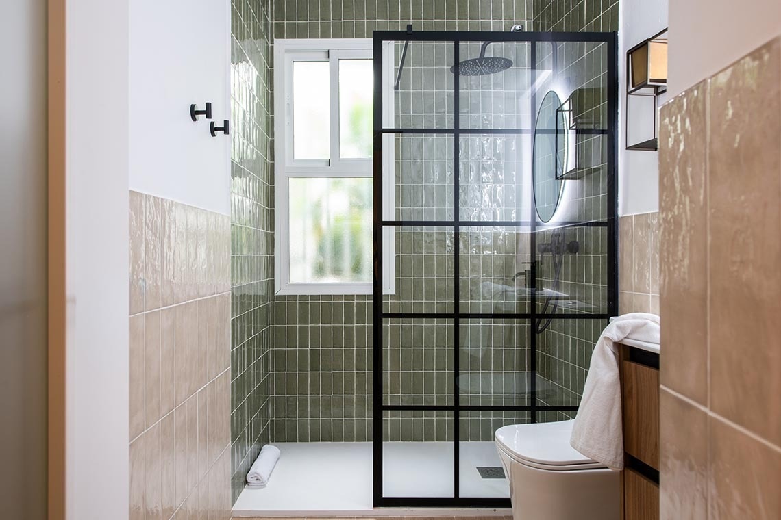 Un moderno cuarto de baño presenta una ducha a ras de suelo con azulejos verdes, una mampara de cristal con marco negro, una ventana y paredes de azulejos beige.