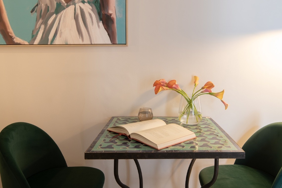 Una mesa de mosaico verde con dos sillas verdes tiene un libro abierto, un vaso y un jarrón de lirios cala naranjas, y arriba una pintura de una figura vestida de blanco.