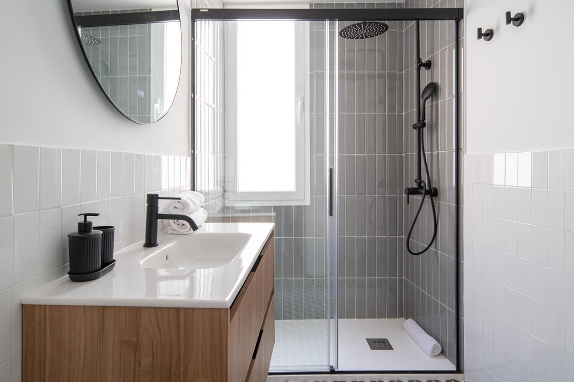 Un baño moderno cuenta con una ducha de mampara y accesorios negros, azulejos grises verticales en la ducha, azulejos blancos horizontales en las paredes, y un mueble de lavabo de madera con una encimera blanca.