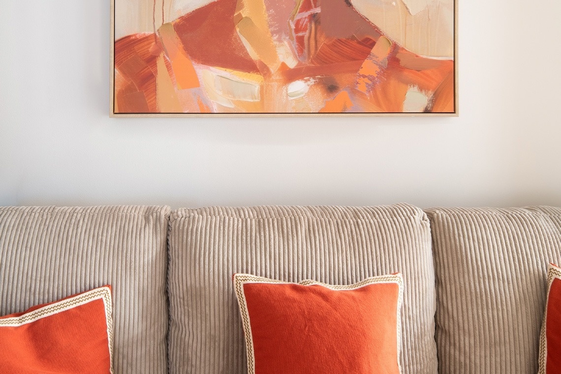 Una pintura abstracta con tonos cálidos cuelga sobre un sofá de pana beige con cojines decorativos naranjas.