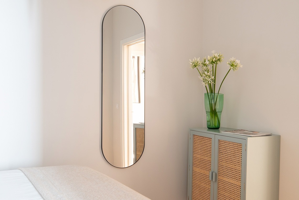Una habitación luminosa presenta un espejo ovalado de cuerpo entero, parte de una cama y un gabinete decorado con un jarrón de flores blancas.