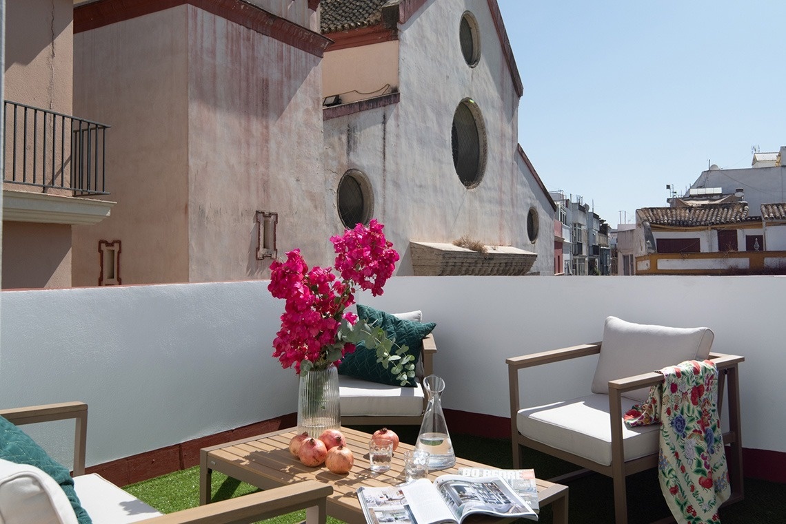 Una acogedora terraza con mobiliario de exterior, flores y granadas, ofrece vistas a edificios antiguos bajo un cielo despejado.