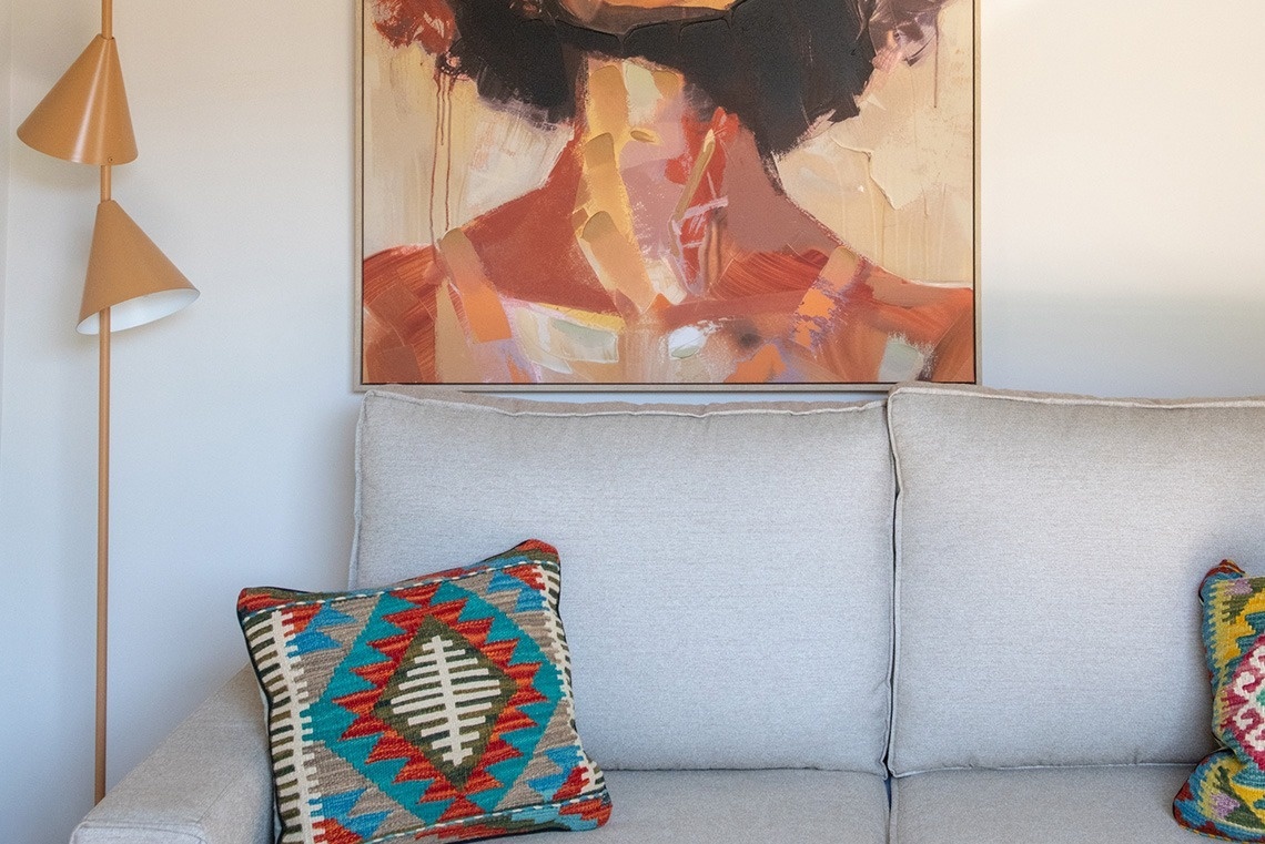 Un sofá de color claro con un cojín de patrones geométricos y coloridos se encuentra debajo de un cuadro abstracto en tonos cálidos, junto a una moderna lámpara de pie con pantallas cónicas.