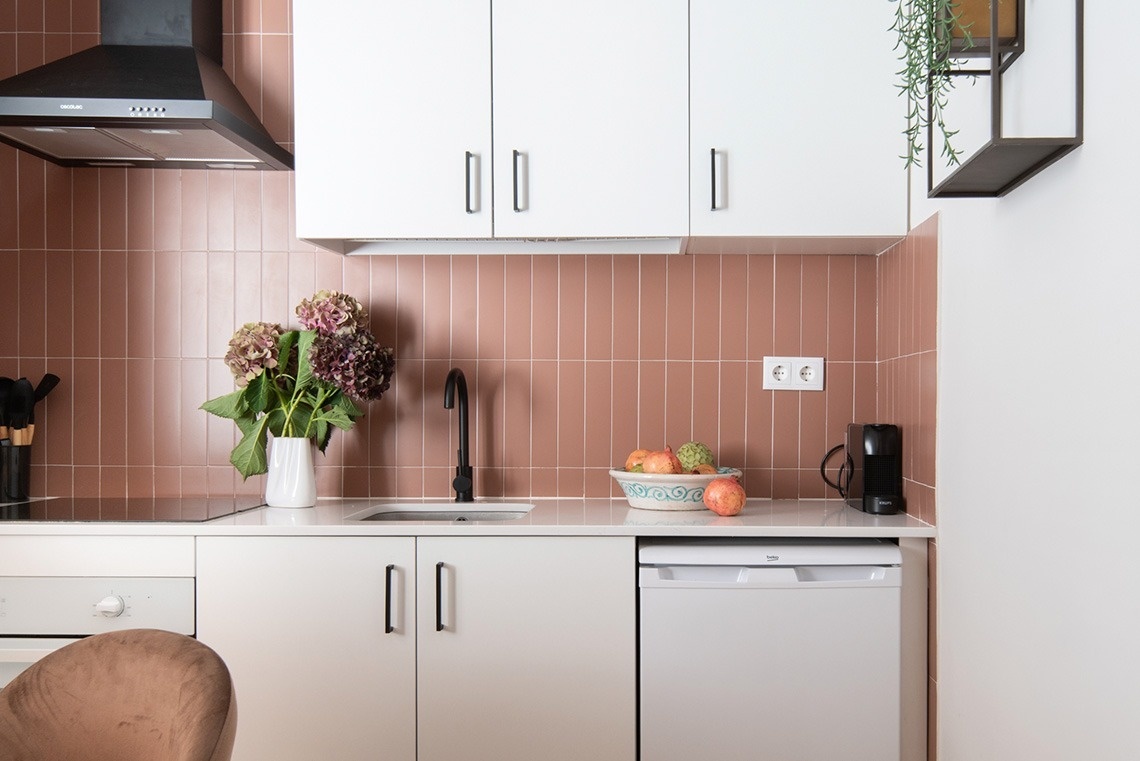 Una cocina moderna presenta un salpicadero de azulejos verticales terracota, gabinetes blancos, encimera clara, y detalles decorativos como flores y un tazón de fruta.