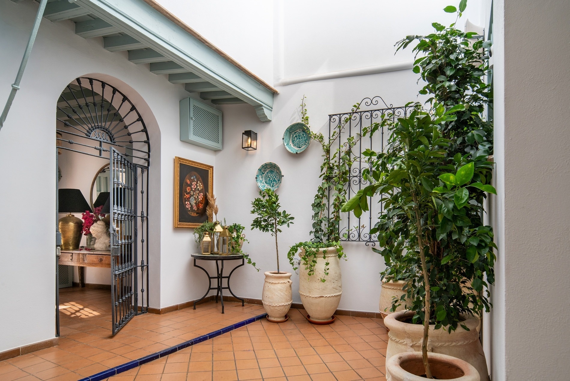 Un luminoso patio interior o pasillo está decorado con plantas, suelo de terracota, una puerta de hierro forjado y acentos en tonos azul verdoso.