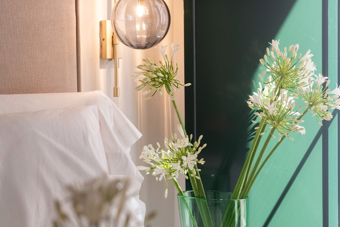 Flores blancas en un jarrón verde decoran un rincón de dormitorio con cabecero claro, ropa de cama blanca y una lámpara de pared dorada, sobre una pared verde oscura con sombras.