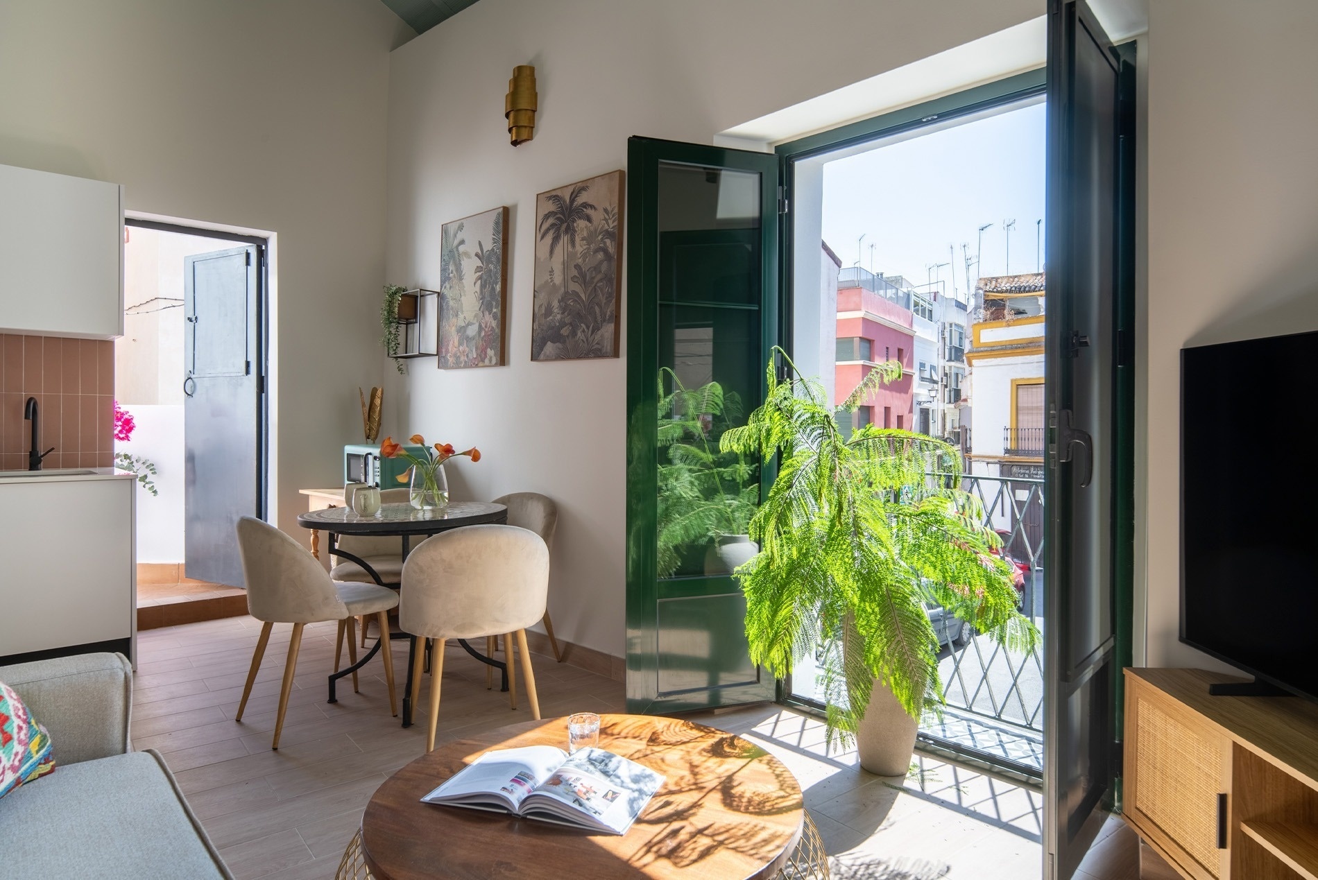 Luminoso interior de apartamento con balcón abierto a una calle urbana soleada, decorado con una gran planta y mobiliario moderno.