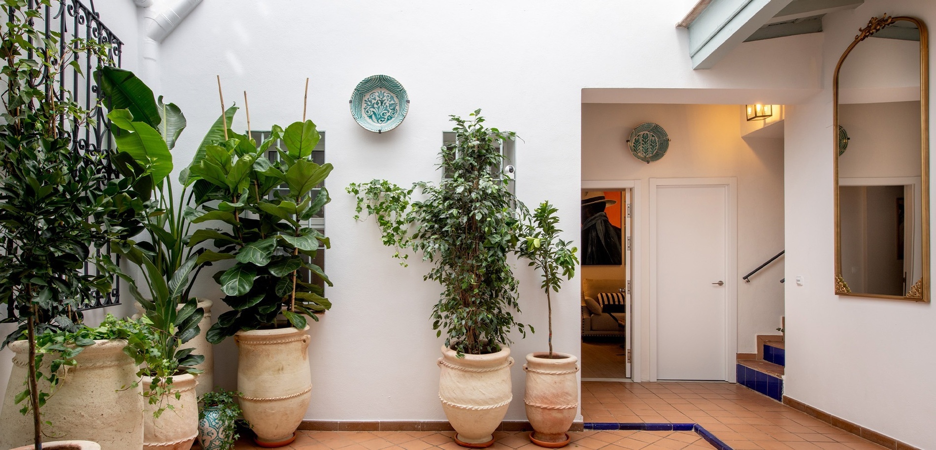 Un luminoso patio interior de paredes blancas y suelo de terracota con detalles azules, decorado con grandes plantas en macetas, un espejo dorado y una entrada que da a unas escaleras y otras habitaciones.