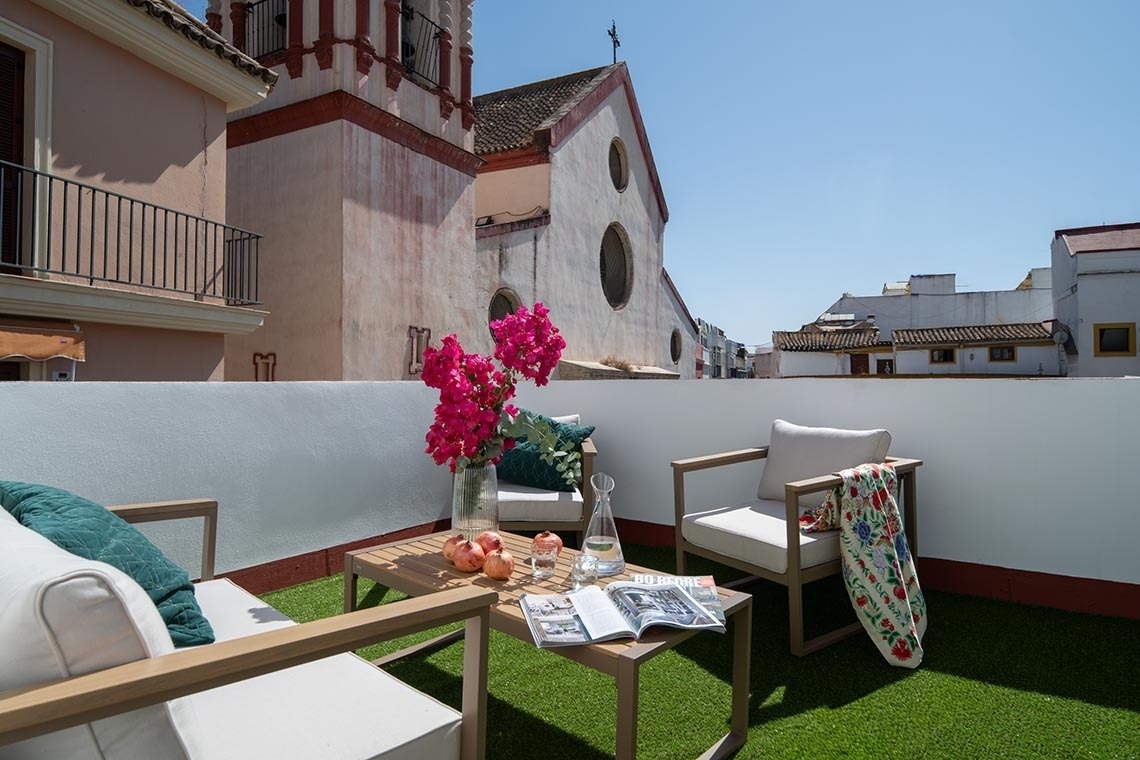 Una azotea soleada con muebles de exterior de madera clara, decorada con flores rosas y granadas sobre césped artificial, ofrece vistas a edificios antiguos y una torre de iglesia bajo un cielo azul claro.
