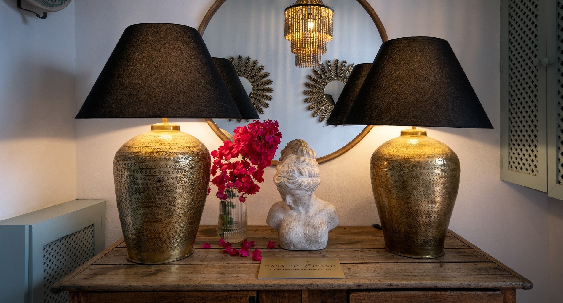 Una elegante composición decorativa presenta dos lámparas doradas con pantallas negras, flores rosadas y un busto sobre una mesa de madera rústica, frente a un gran espejo circular.