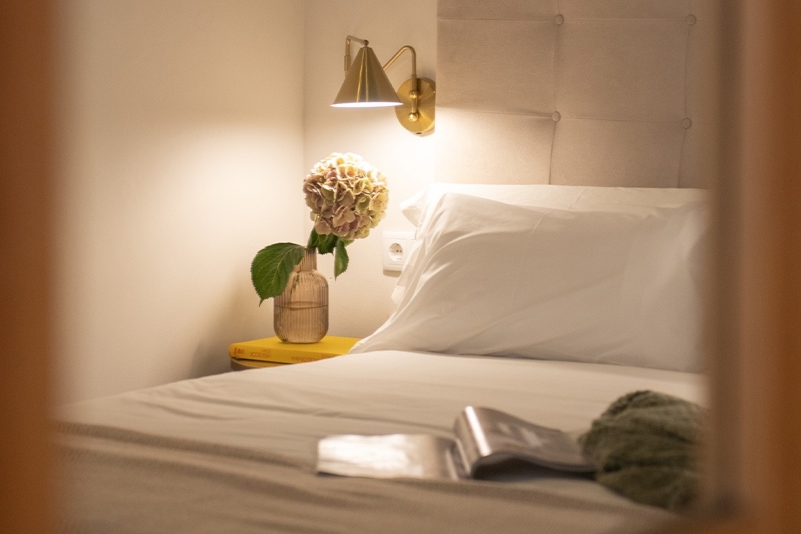 Primer plano de una cama acogedora con una mesita de noche decorada con hortensias y libros, bajo la cálida luz de una lámpara de pared dorada.