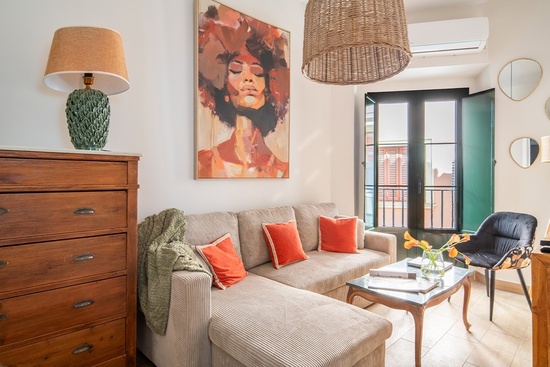 Se muestra un salón acogedor y bien iluminado, con un sofá seccional de color neutro, cojines naranjas, una cómoda de madera, un cuadro vibrante de una mujer y una ventana con contraventanas verdes.