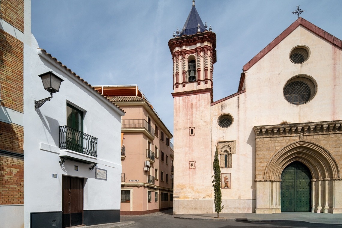 La imagen muestra una calle de pueblo con una iglesia de fachada clara y un campanario distintivo, junto a edificios residenciales de estilo tradicional.
