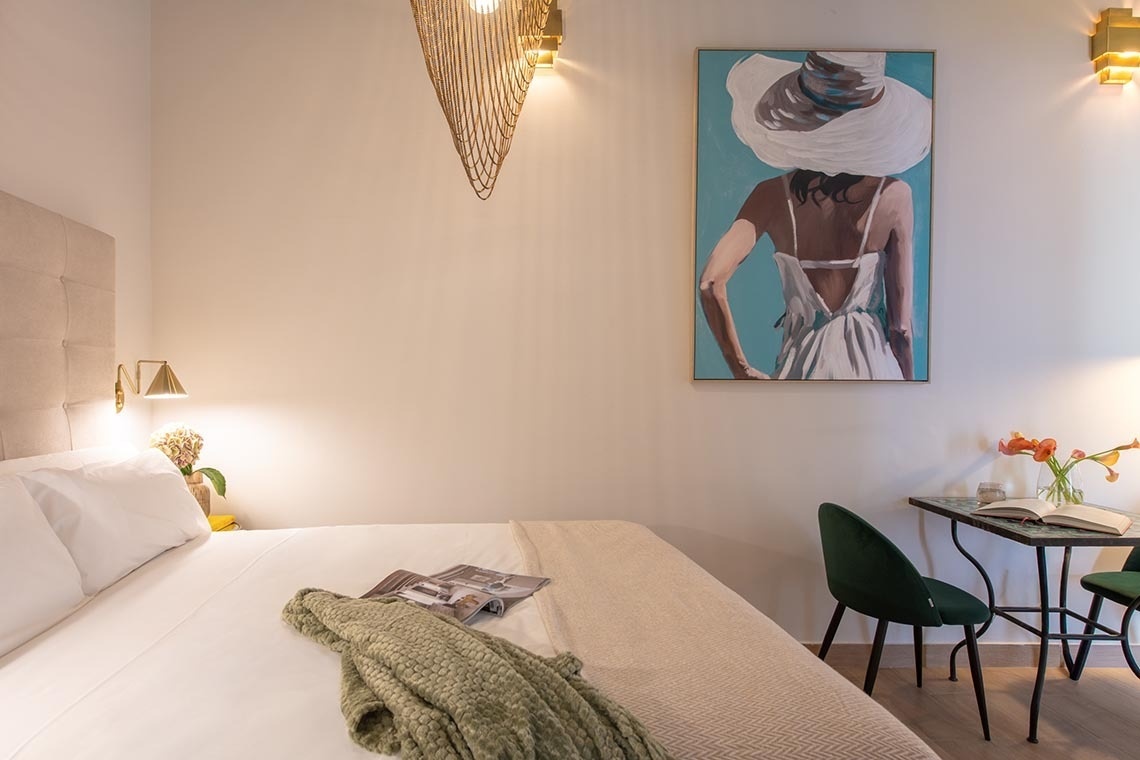 Un dormitorio luminoso presenta una cama con ropa blanca, un cuadro de una mujer con sombrero blanco en la pared y un pequeño rincón de mesa con dos sillas verdes.