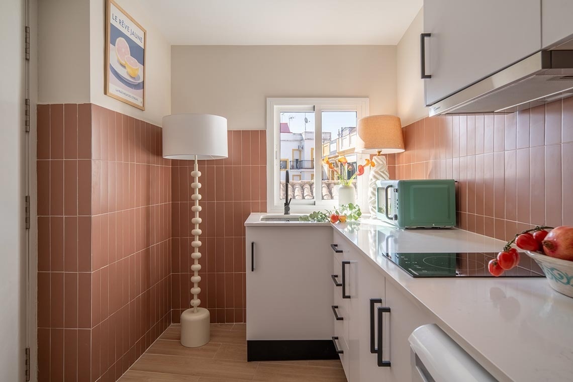 Una cocina moderna y acogedora con azulejos verticales color terracota, muebles blancos y una ventana que da a la calle.