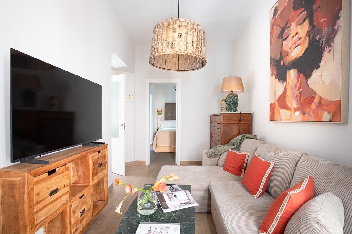 Una sala de estar luminosa y acogedora presenta un sofá seccional con cojines rojos, un televisor sobre un mueble de madera, una mesa de centro con flores y una lámpara de mimbre, con una pintura de una mujer en la pared.