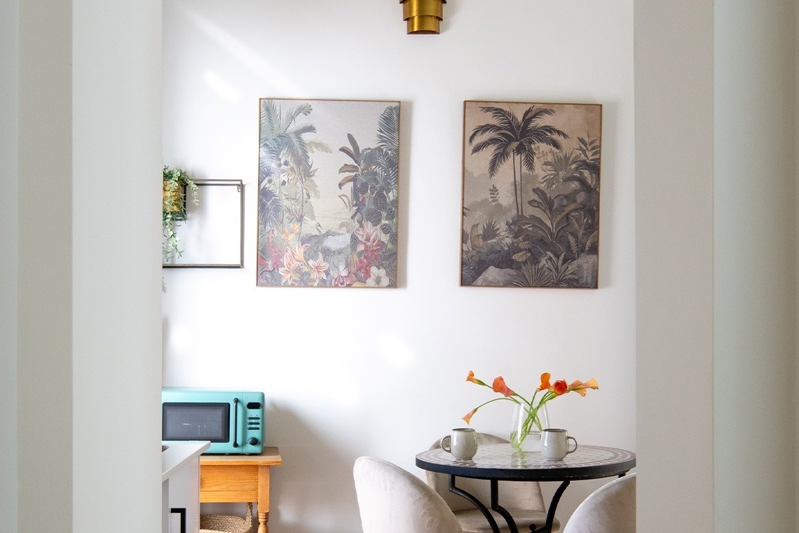 La imagen muestra una acogedora área de comedor decorada con dos cuadros de temática tropical, una mesa redonda con flores y tazas, y un microondas turquesa en un rincón.