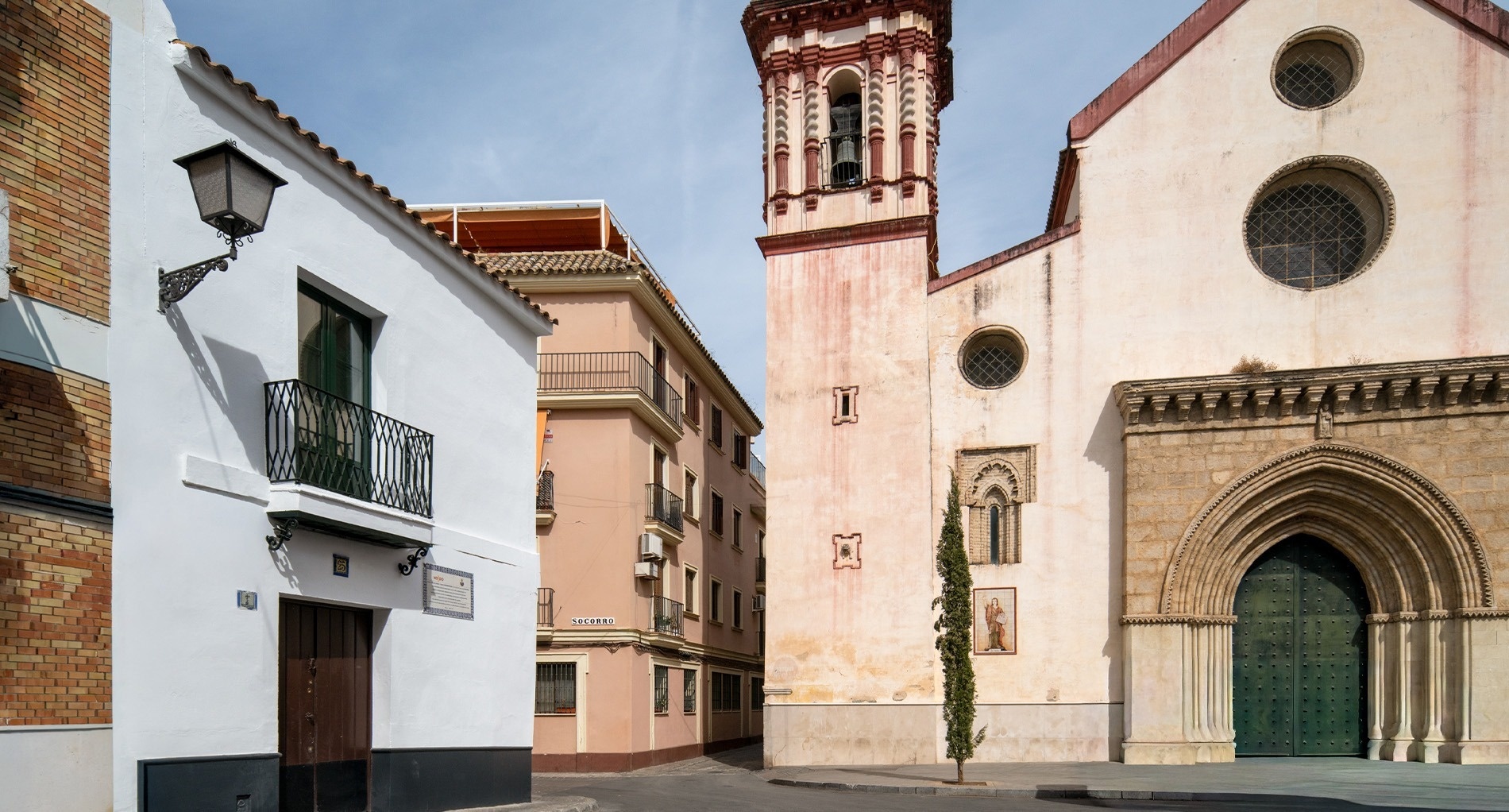 Una calle en un día soleado muestra una iglesia de fachada clara con un campanario y una gran entrada arqueada, junto a una casa blanca con balcón y farola, y edificios de color rosa al fondo.