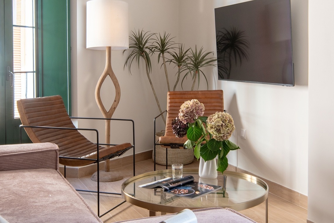 Un salón moderno y luminoso cuenta con un sillón de cuero, una lámpara de pie escultural de madera, plantas decorativas y una mesa de centro de cristal con un jarrón de hortensias.