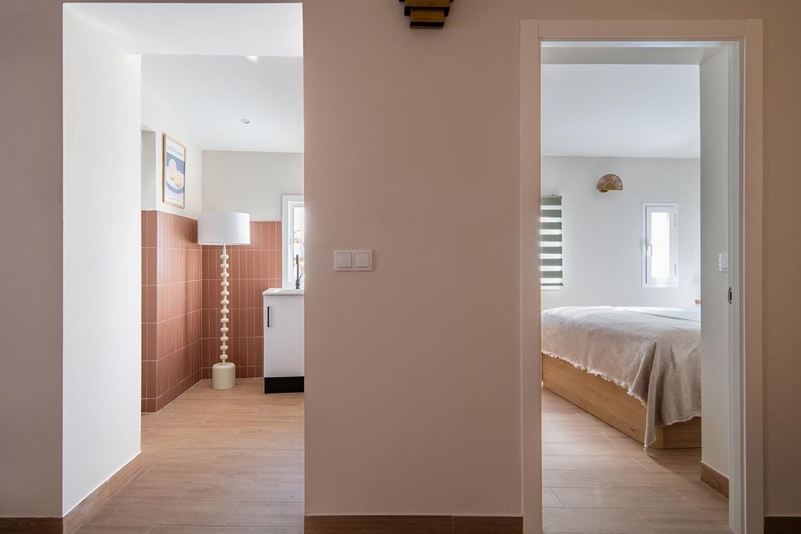 La imagen muestra un pasillo iluminado que conecta dos habitaciones diferentes, una con paredes de azulejos terracota y una lámpara, y otra con una cama y ventanas.