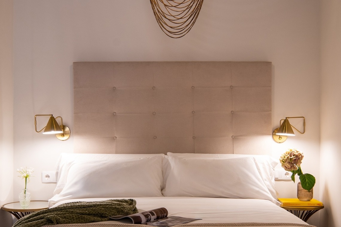 Una cama con cabecero acolchado está flanqueada por apliques dorados y mesitas con flores, con una decoración de cadenas doradas colgando sobre ella.