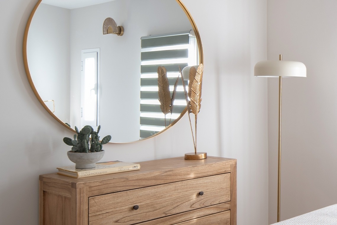 Una cómoda de madera con un espejo redondo dorado, un cactus en maceta y adornos de plumas doradas, junto a una lámpara de pie en un interior moderno.