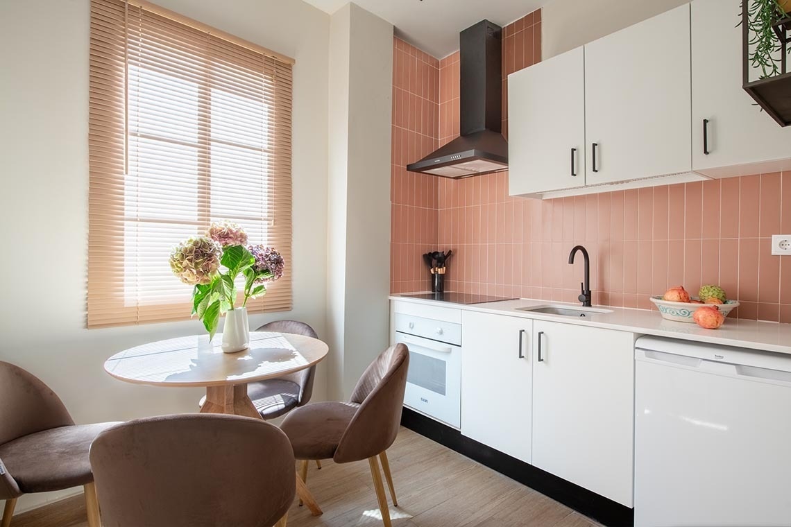Una cocina moderna y luminosa cuenta con una pared de azulejos rosas, gabinetes blancos, una campana extractora negra y una mesa redonda de madera con sillas marrones, adornada con un jarrón de flores junto a una ventana con persianas.