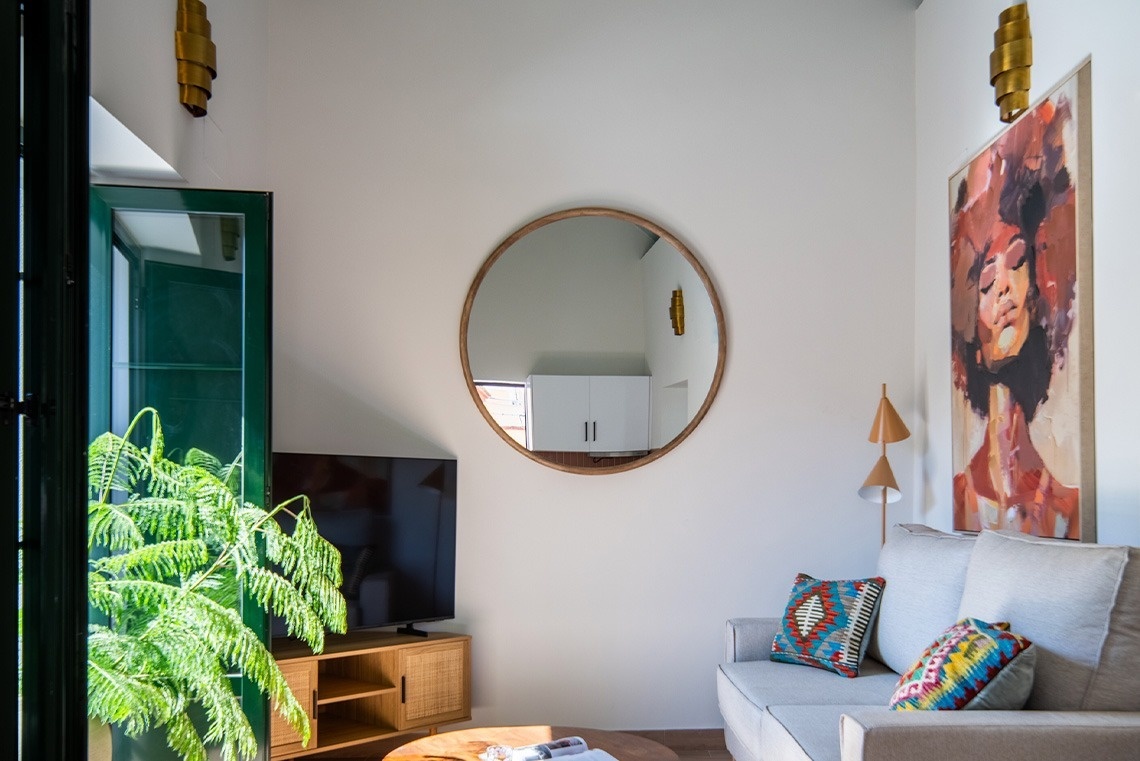 Una luminosa sala de estar con un sofá gris, un televisor, una planta verde vibrante, un espejo redondo en la pared y un colorido cuadro de una mujer.