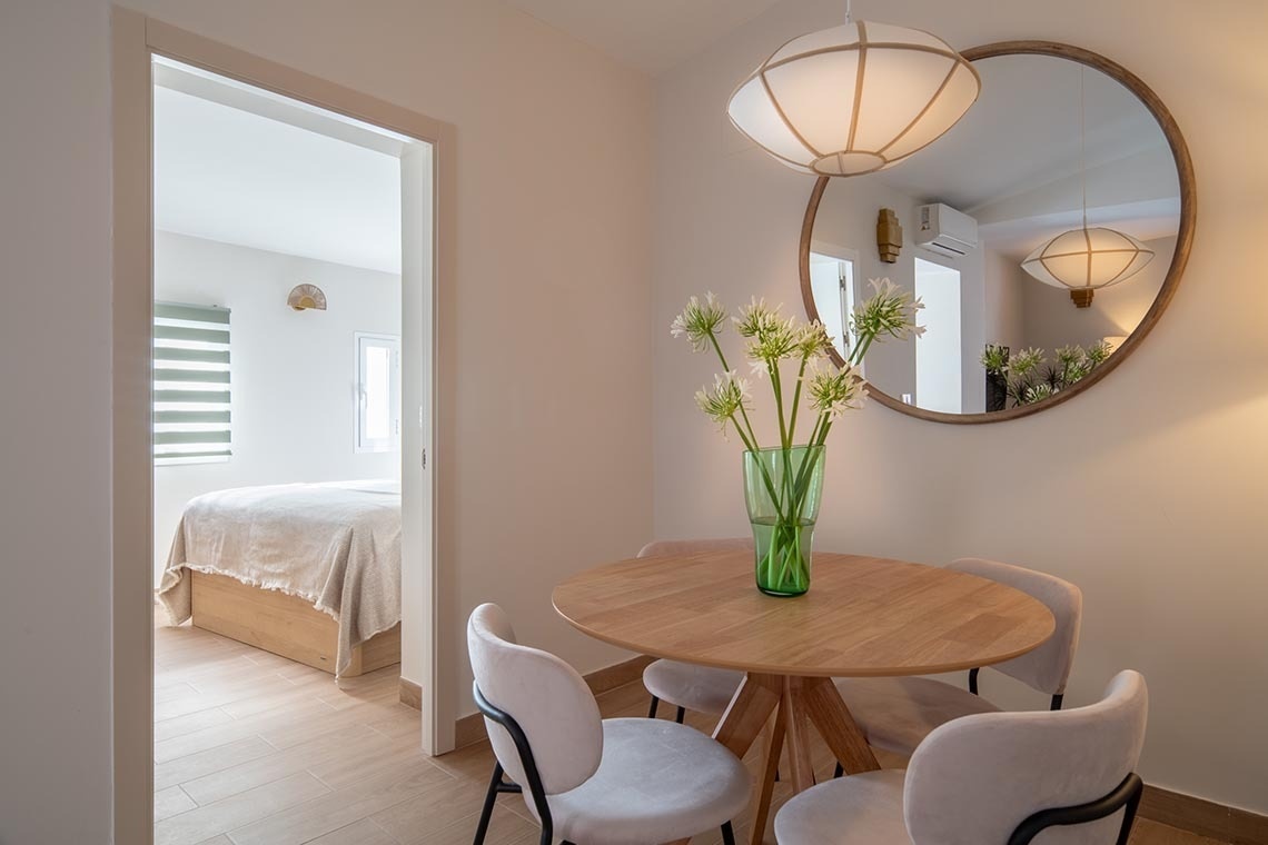 Se muestra un comedor luminoso con una mesa redonda de madera, sillas tapizadas y un jarrón de flores, con un gran espejo circular en la pared y un dormitorio visible al fondo a través de una puerta.