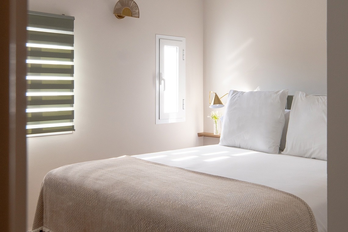 Un luminoso dormitorio de tonos neutros presenta una cama con sábanas blancas y una manta beige, dos ventanas (una con persiana) y una lámpara de pared dorada, todo bañado por la luz del sol.