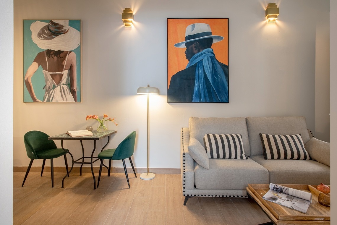 Un salón moderno y acogedor con arte figurativo en las paredes, un sofá claro, una pequeña mesa con sillas verdes y una iluminación cálida.