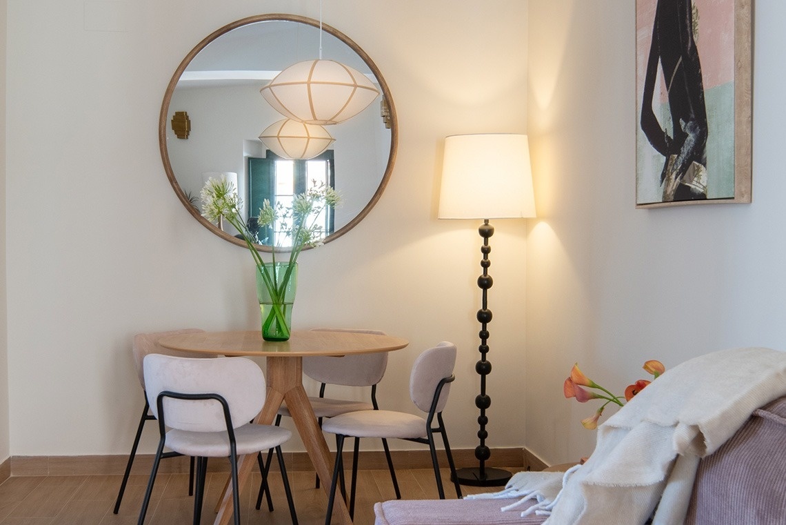 Una escena interior luminosa muestra un rincón de comedor con una mesa redonda de madera, sillas modernas, un espejo circular decorativo y un jarrón con flores blancas, complementado por una lámpara de pie en la esquina.