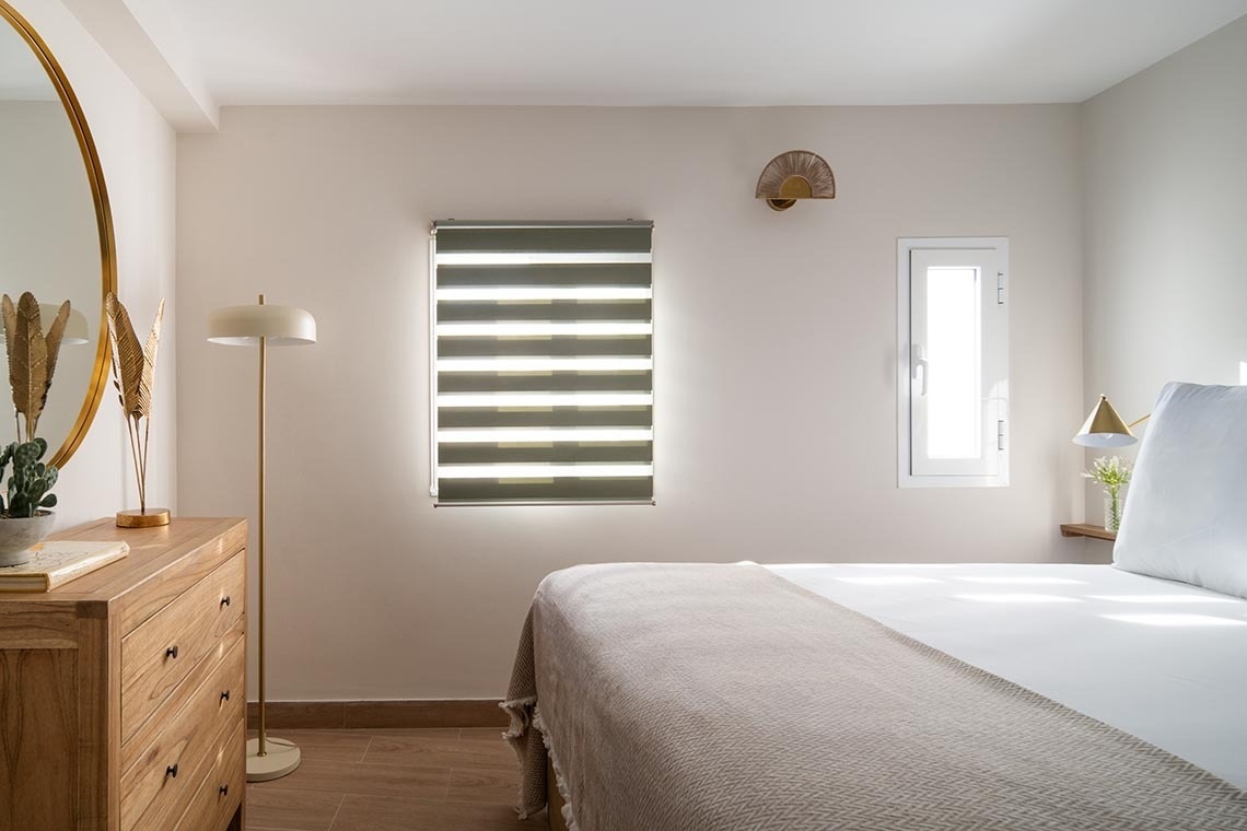 Un dormitorio moderno y sereno en tonos neutros, con una cama de ropa blanca, una cómoda de madera junto a un espejo redondo, y dos ventanas, una equipada con una persiana tipo cebra.