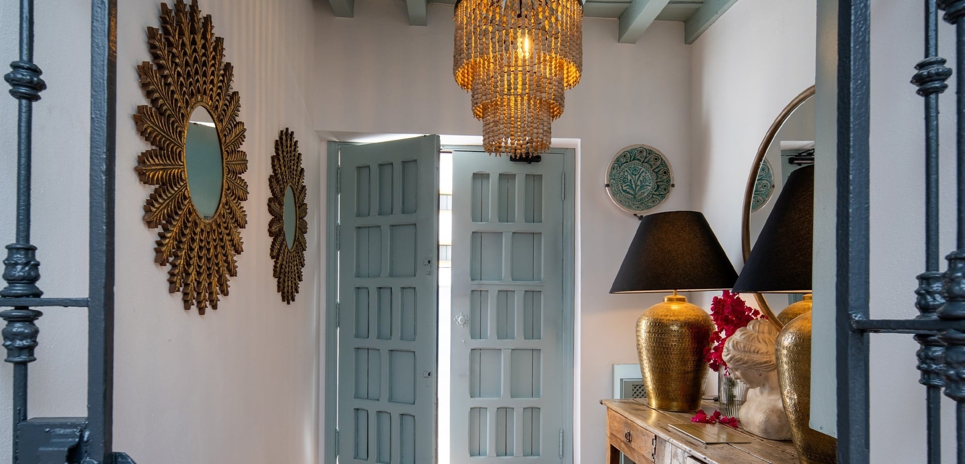 Un pasillo elegantemente decorado presenta espejos dorados en forma de sol, puertas azules parcialmente abiertas, una lámpara de araña de cuentas de madera, y una mesa con lámparas, un espejo y adornos.