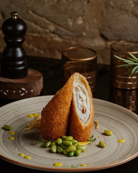 Un rollo empanizado y frito, cortado por la mitad para mostrar su cremoso relleno, se presenta verticalmente en un plato con edamame y salsa amarilla, con elementos de cocina en el fondo.