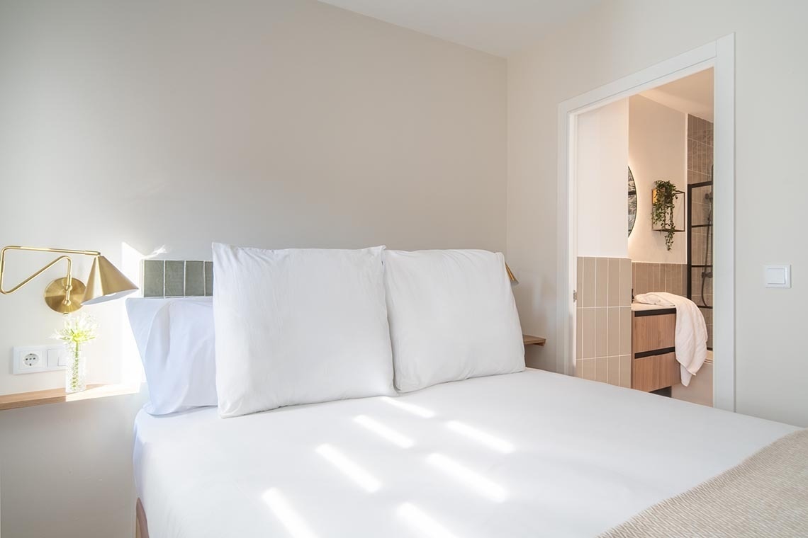 Un dormitorio luminoso con una cama blanca, una lámpara de pared dorada y acceso a un baño moderno con ducha.