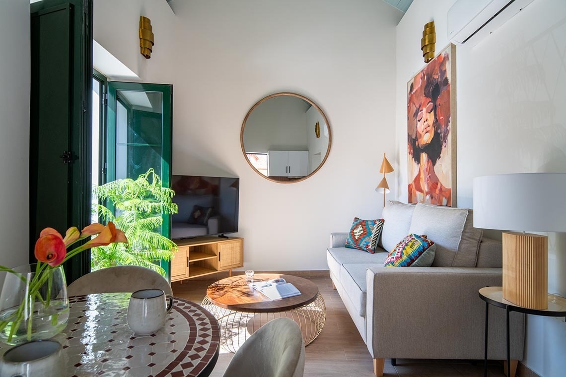 Un salón moderno y luminoso decorado con un sofá, mesa de centro, un espejo redondo, un vibrante cuadro, una mesa de comedor con mosaico y ventanas con contraventanas verdes que dan al exterior.